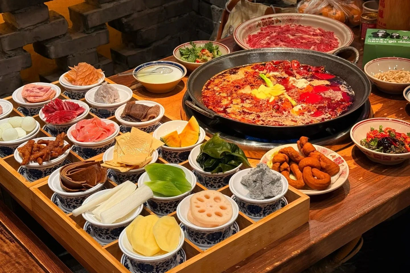 Sichuan hot pot bubbling with chili and peppercorns, surrounded by small plates of meat, vegetables and tofu ready to cook