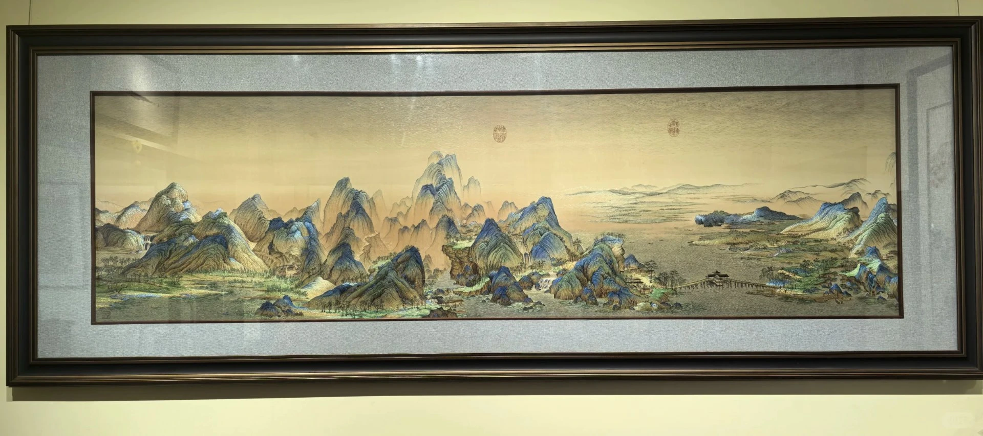 Su embroidery (苏绣) landscape scroll — Suzhou's signature needlework, displayed in a museum: green-and-blue mountain ranges with mist, traditional Chinese ink-wash painting recreated in silk thread